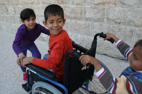 Niño usuario de silla de ruedas sonríe a un niño que lo va empujando y delante de la silla una niña sujeta la silla, se perciben divertidos.
