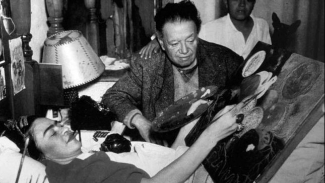 Frida Kahlo, pinta en un caballete adaptado mientras convalece en su cama, a un lado Diego Rivera observa.