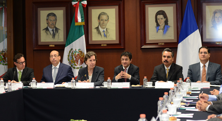 Funcionarios en reunión sentados alrededor de una mesa y a sus espaldas cuadros de otros funcionarios, y las banderas de México y Francia 