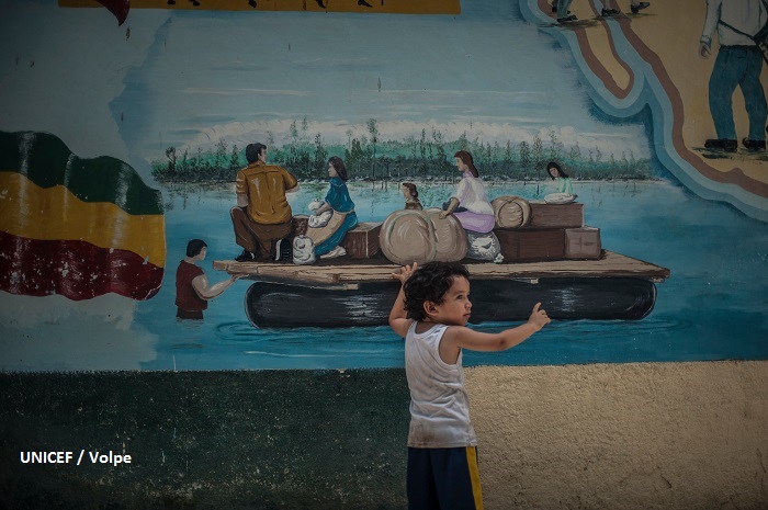 Niño migrante en casa de refugio. Al fondo pintura mural naif con una familia de migrantes que cruza el río.
