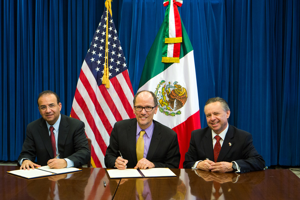 Tres hombres sentados frente a una mesa, dos de ellos firmando un documento. Los tres sonriendo, detrás de ellos las banderas de Estados Unidos y México.