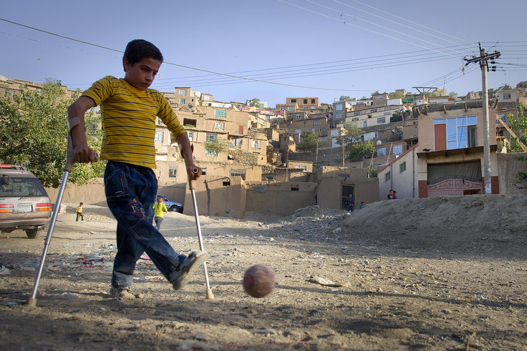 Niño con secuela de polio patea un balón de fútbol, apoyado en sus muletas canadienses