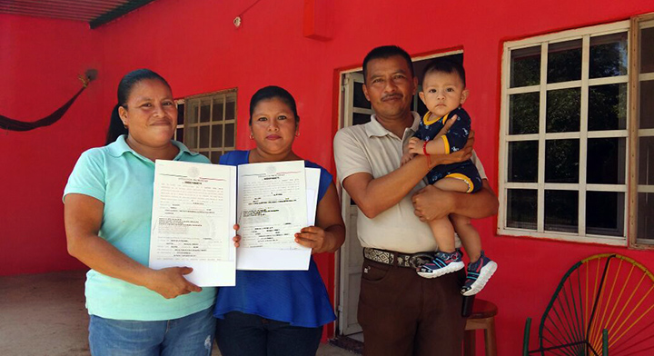 Familia con documentos agrarios en la mano, y de fondo casa roja