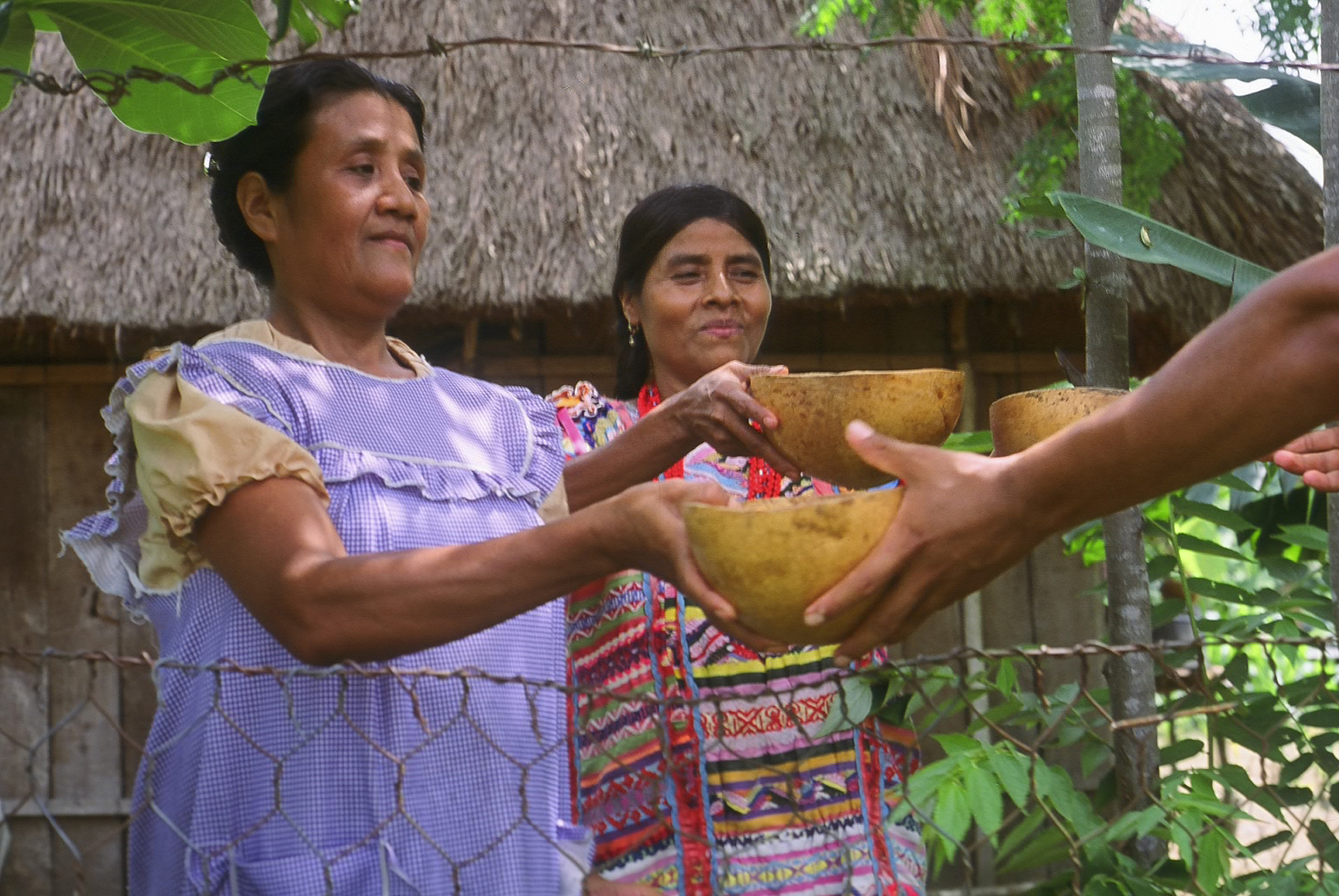 Vista general de dos mujeres indígenas dando recipientes-bandejas en localidad natural.