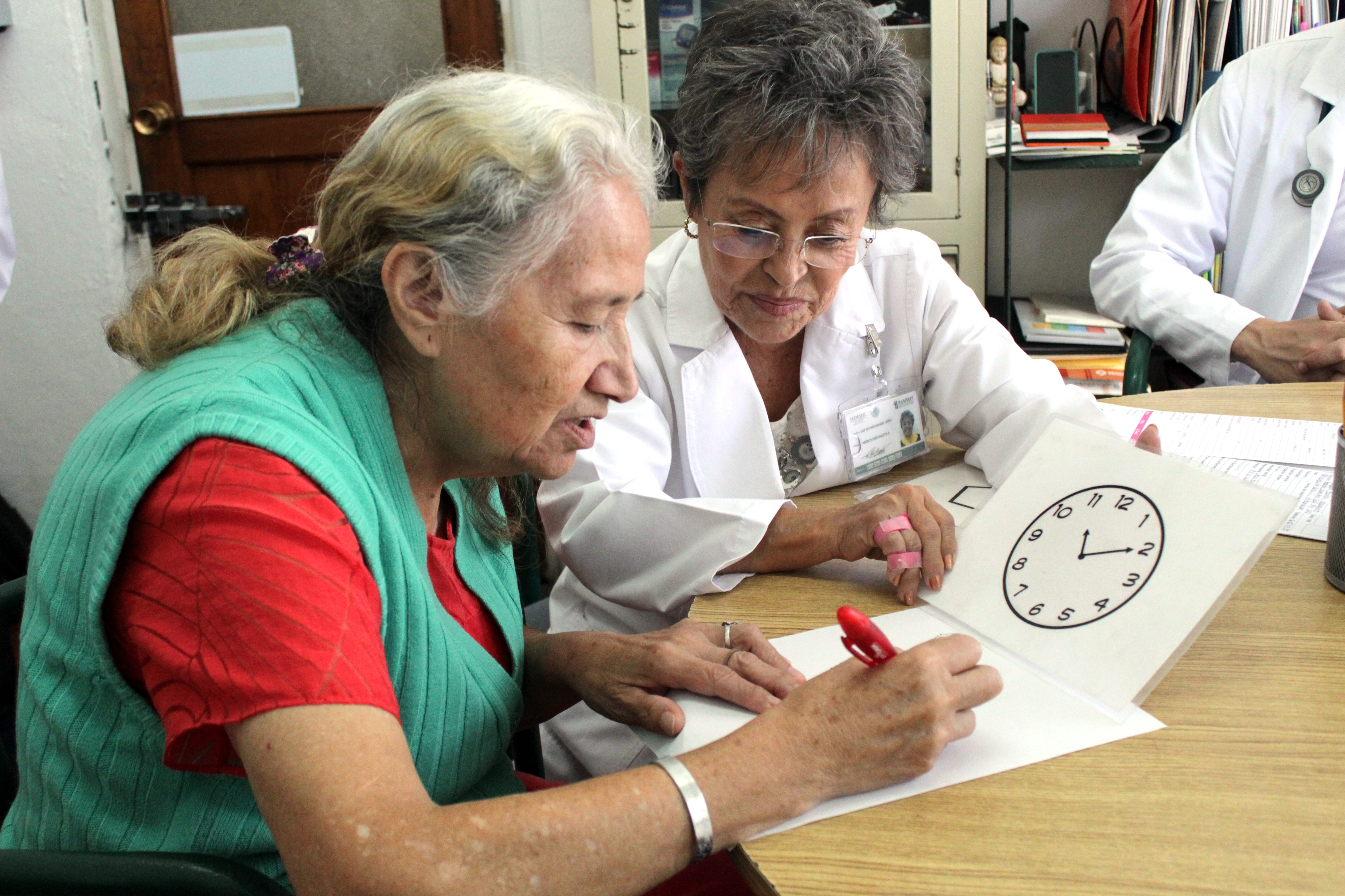 Una doctora y una adulta mayor, la adulta mayor empieza a dibujar un reloj. 