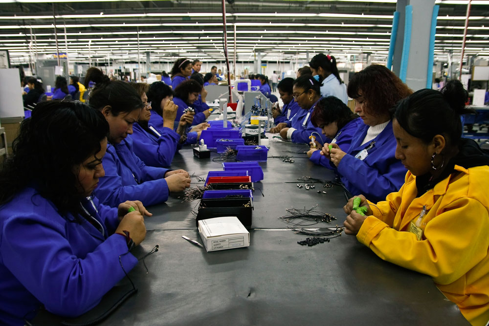 Varias mujeres trabajando en una fabrica. Traen uniforme azul y amarillo están de ambos lados de una mesa de trabajo.