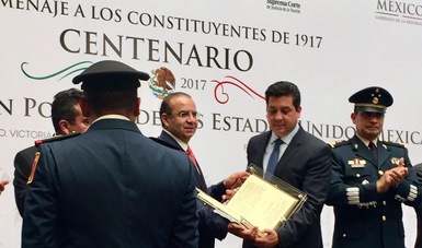 Dos personas de pie, mostrando un facsímil de la Constitución Política Mexicana, rodeados de dos miembros del ejército.