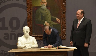 Dos personas de pie una de ellas firmando un documento y detrás una fotografía de Venustiano Carranza