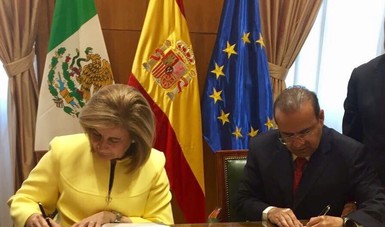 Dos personas, un hombre y una mujer firmando un documento, las banderas de España, México y la Unión Europea detrás de ellos.