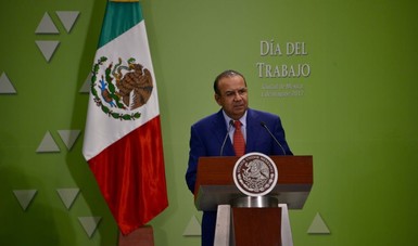 Un hombre hablando al micrófono, detrás de él la bandera de México.