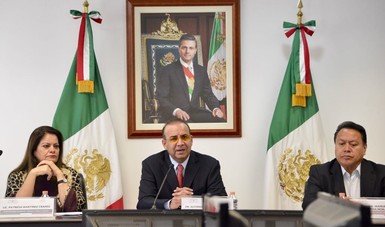 Tres personas sentadas en una mesa, detrás de ellos una fotografía del Presidente de la República y dos banderas de México.