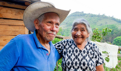 Dos adultos mayores sonriendo. de fondo un paisaje verde y su choza.