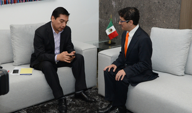 Jorge Ramos entrevistando al Secretario Virgilio Andrade, se les ve sentados en una sala de color blanco y en una mesa se ve la bandera de México 