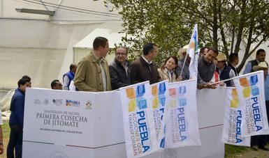 Varias personas observando el banderazo de salida que da el Secretario del Trabajo, detrás de una valla blanca, portando banderas que dicen Puebla.
