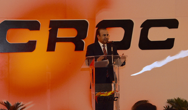 Un hombre de pie hablando al micrófono, detrás de él el en un fondo naranja con letras negras, las siglas de la CROC.