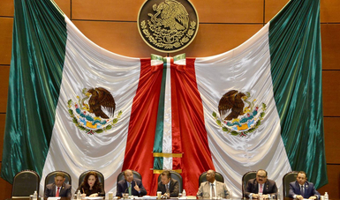 Siete personas sentadas a una mesa, detrás de ellos dos banderas de México flanqueando a cada lado.