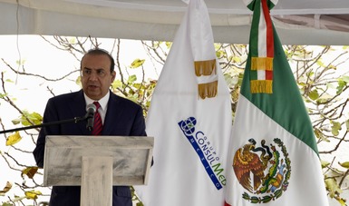 Un hombre de pie hablando al público, a su lado la bandera de México.